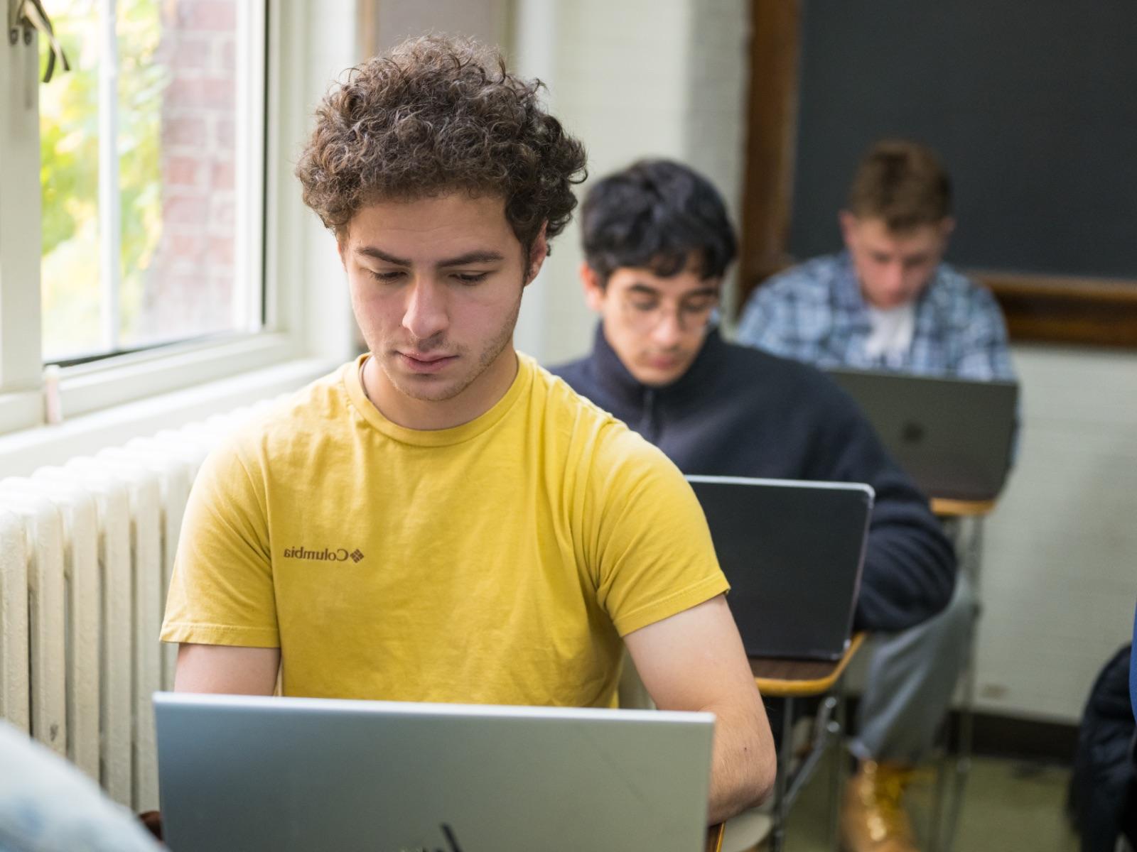 Three students sit at desks one in front of another in a classroom while looking at their laptops